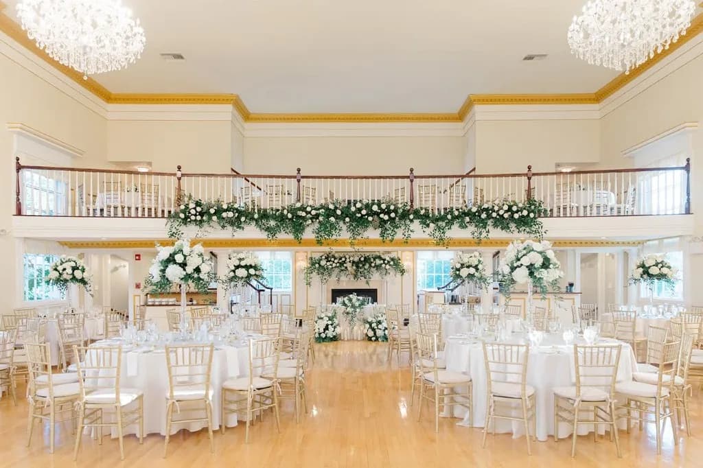 Grand ballroom wide view — both crystal chandeliers, balcony with garland, gold Chiavari chairs, tall centerpieces at The Commons 1854